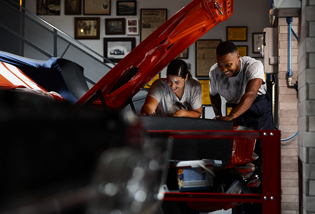 Two mechanics smiling while working under a car hood in a garage, surrounded by tools and framed pictures on the wall.