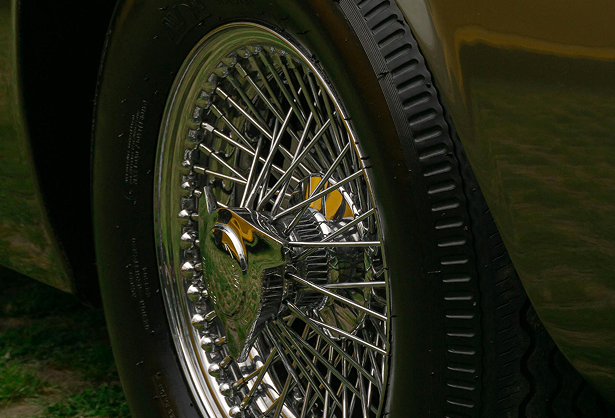 Close-up of a classic car's chrome wire-spoke wheel and tire on grass, reflecting surrounding greenery.